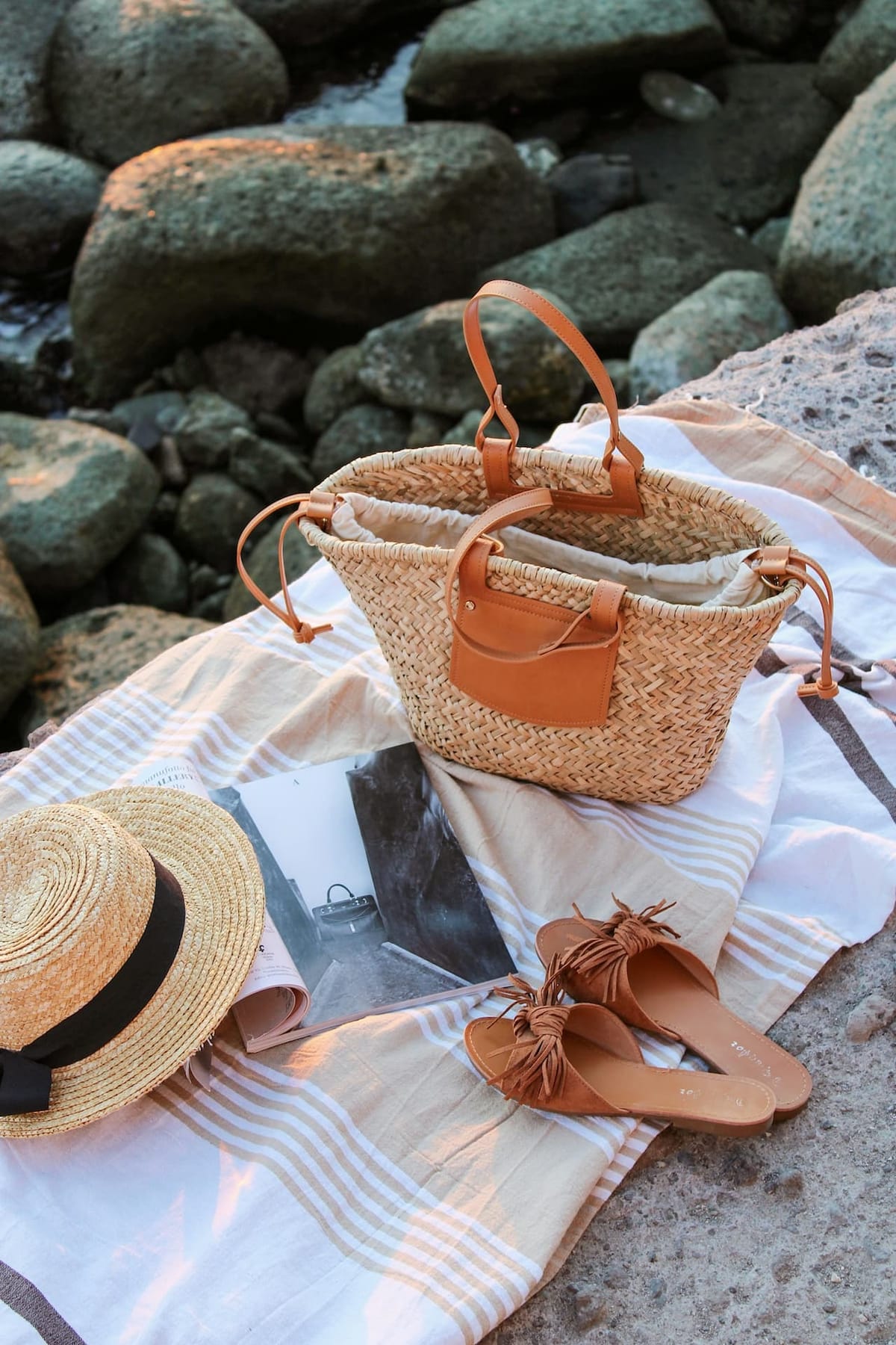 Sac cabas en raphia posé sur une serviette de plage avec chapeau de paille et sandales – accessoire mode bohème chic pour l’été 2025.