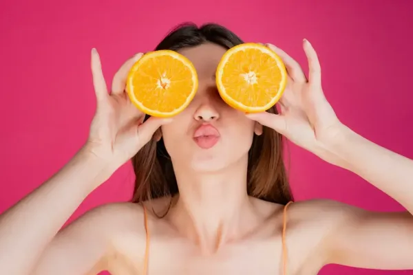 Femme souriante tenant deux demi-oranges devant ses yeux, fond rose vif et expression amusée.